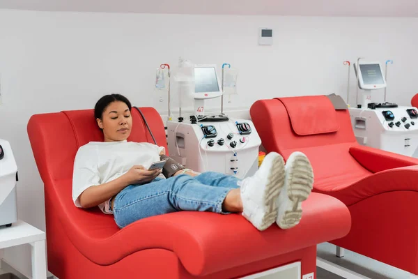 Donna multirazziale navigazione internet su smartphone mentre seduto su comoda sedia medica vicino alla macchina trasfusione mentre donare il sangue in clinica, procedura medica — Foto stock