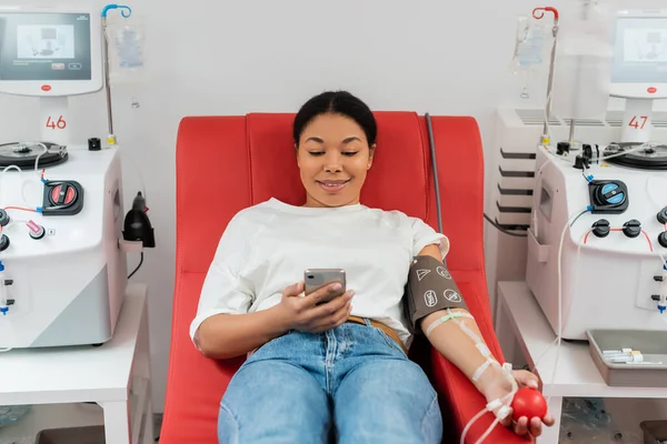 Lächelnde multirassische Frau in Bluttransfusionsset mit Gummiball und Internet-Surfen auf dem Handy, während sie auf einem medizinischen Stuhl in der Nähe automatisierter Geräte im Labor sitzt — Stockfoto