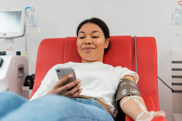 Pleased multiracial woman in blood pressure cuff messaging on mobile phone while sitting on medical chair near automated transfusion machine in modern laboratory, medical procedure — Stock Photo