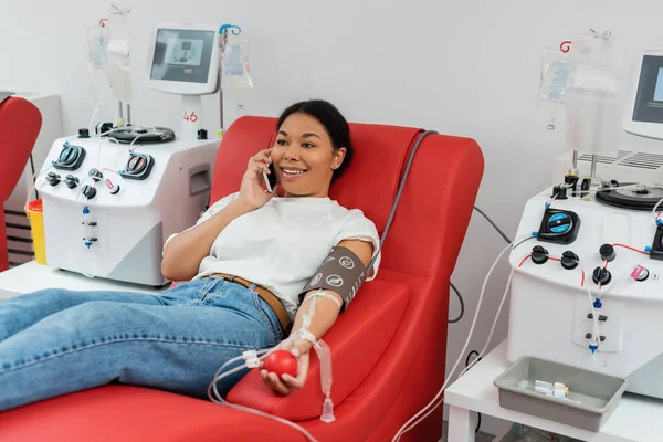 Счастливая многорасовая женщина с резиновым мячом сидит на медицинском стуле и разговаривает по мобильному телефону возле аппаратов для переливания крови во время сдачи крови в лаборатории — стоковое фото