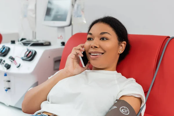 Junge und fröhliche multirassische Frau, die auf dem Smartphone spricht, während sie auf dem Behandlungsstuhl in der Nähe eines verschwommenen Transfusionsgeräts im Blutspendezentrum sitzt — Stockfoto