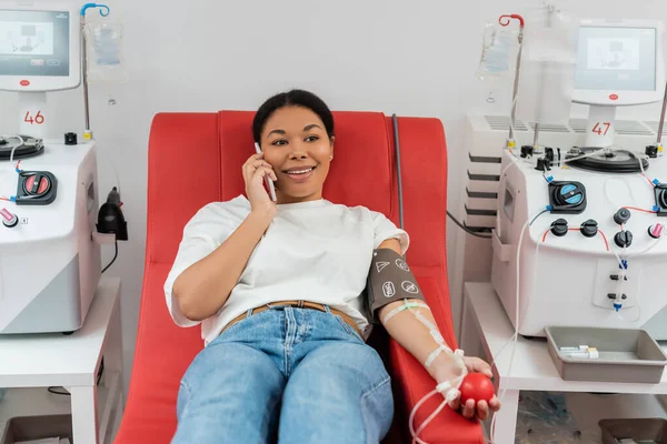 Femme souriante multiraciale avec manchette de pression et boule de caoutchouc assise sur une chaise médicale près des machines à transfusion et parlant sur un téléphone portable pendant le don de sang à la clinique — Photo de stock