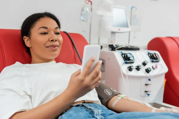 Задоволена багаторасова жінка сидить на зручному медичному стільці і обмін повідомленнями на мобільному телефоні біля автоматизованої машини переливання крові в лабораторії, розмитий фон — стокове фото