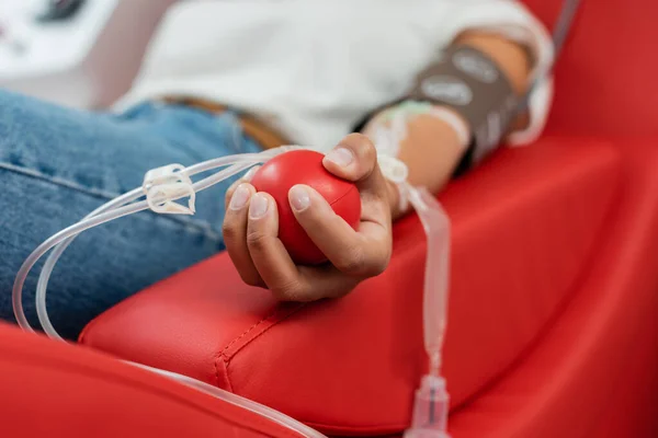 Enfoque selectivo de la bola de goma en la mano de la mujer multirracial con transfusión conjunto sentado en la silla médica cómoda y la donación de sangre en el laboratorio - foto de stock