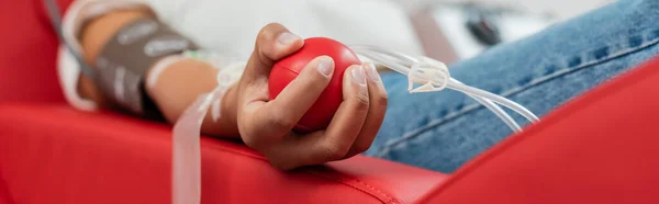 Частичный взгляд на многорасовую женщину с набором для переливания крови, держащую резиновый мяч, сидя на эргономичном медицинском стуле во время сдачи крови в лаборатории, баннер — стоковое фото