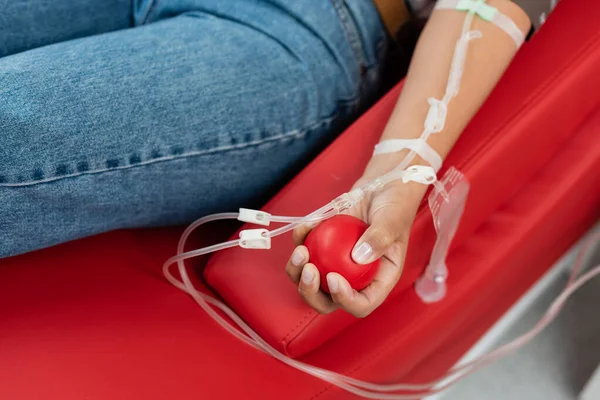 Partial view of multiracial woman with a transfusion set holding rubber ball while sitting on ergonomic medical chair during blood donation in clinic, medical procedure — Stock Photo