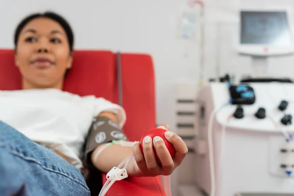 Селективный фокус медицинского резинового мяча в руке многорасовой женщины, сидящей на удобном медицинском стуле возле машины для переливания крови при сдаче крови в лаборатории, размытый фон — стоковое фото