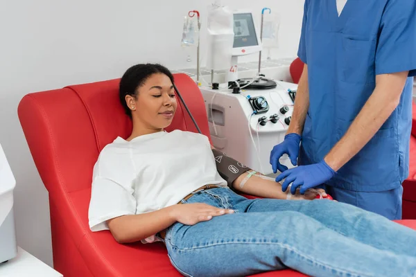 Медицинский работник в синей форме и латексных перчатках регулирует переливание крови, установленное рядом с многорасовой женщиной, сидящей на медицинском стуле и сдающей кровь в клинике, медицинская процедура — стоковое фото