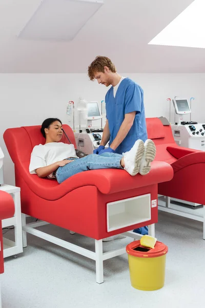 Médico pelirrojo en uniforme azul y guantes de látex cerca de máquinas de transfusión automatizadas y mujer multirracial en silla médica donando sangre en el hospital, procedimiento médico - foto de stock