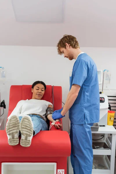 Pelirroja trabajadora de la salud en uniforme azul ajustando transfusión fijada en mujer multirracial sentada con pelota de goma en silla médica mientras dona sangre en clínica - foto de stock