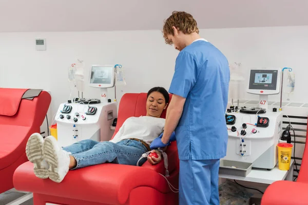 Young redhead doctor in blue uniform adjusting transfusion set on multiracial woman holding rubber ball while sitting on medical chair near transfusion machine in blood donation center — Stock Photo