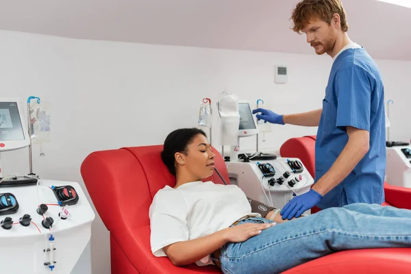 Médico pelirrojo en uniforme azul y guantes de látex operando la máquina de transfusión cerca de la mujer multirracial con manguito de presión sentado en la silla médica en el centro de donación de sangre - foto de stock