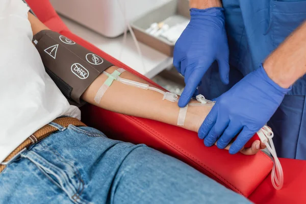 Visão recortada de médico em luvas de látex colando band-aid no braço de mulher multirracial com manguito de pressão e transfusão definido durante doação de sangue na clínica — Fotografia de Stock