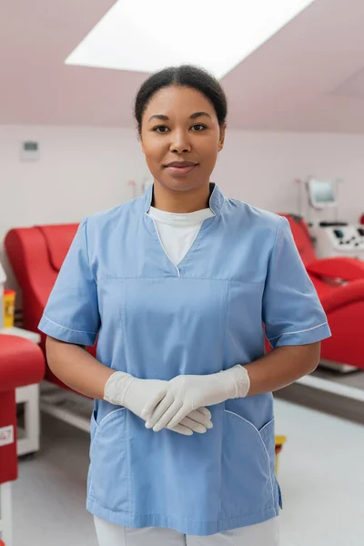 Positive multiracial nurse in blue uniform and latex gloves looking at camera near medical chairs and transfusion machines in blood donation center on blurred background — Stock Photo