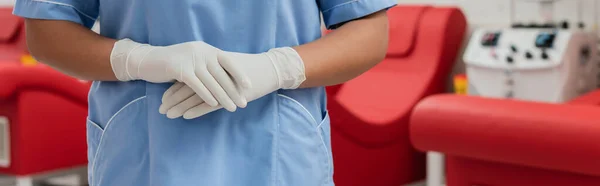 Vista recortada del trabajador sanitario multirracial en uniforme azul y guantes de látex de pie en ambiente estéril del centro de transfusión de sangre, pancarta - foto de stock