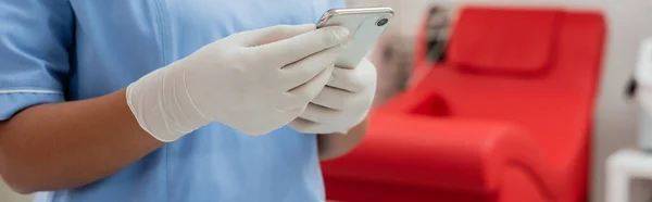 Vista parcial del trabajador sanitario multirracial en uniforme azul y guantes de látex mensajería en el teléfono inteligente cerca de la silla médica borrosa en la estación de transfusión de sangre, pancarta - foto de stock