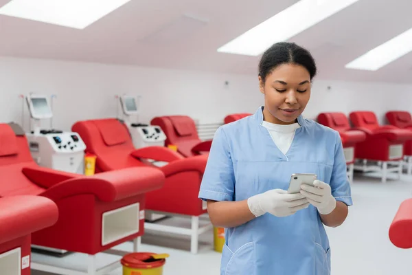 Positive multiracial nurse in blue uniform and latex gloves texting on smartphone near blurred medical chairs and transfusion machines in blood donation center — Stock Photo