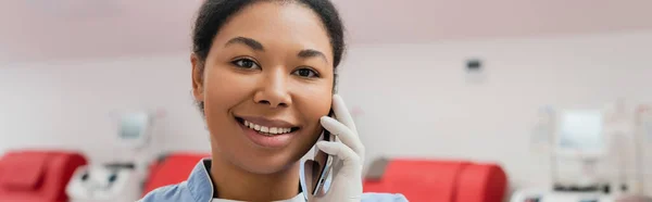 Joven y feliz trabajador de la salud multirracial en guante de látex estéril sonriendo durante la conversación en el teléfono móvil en la estación de transfusión de sangre, pancarta - foto de stock