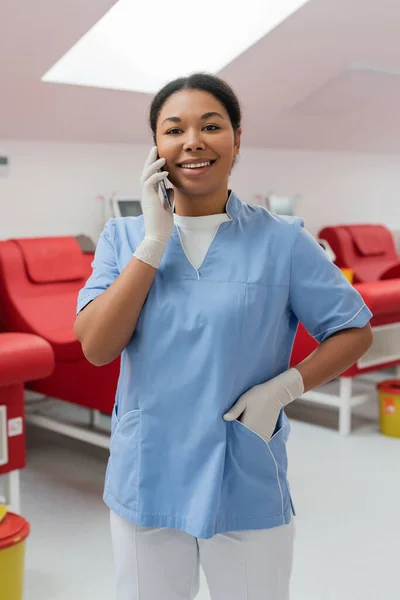Joyeuse infirmière multiraciale en gants de latex debout avec la main dans la poche d'uniforme bleu et parlant sur smartphone près de chaises médicales floues en laboratoire — Photo de stock