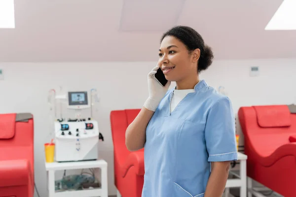 Trabajador sanitario multirracial feliz en uniforme azul y guante de látex hablando en el teléfono móvil cerca de la máquina de transfusión borrosa y sillas médicas en el centro de donación de sangre - foto de stock
