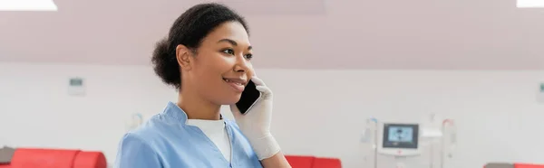 Щасливий багаторасовий медичний працівник у блакитній формі посміхається під час розмови на мобільному телефоні в центрі донорства крові на розмитому фоні, банер — стокове фото
