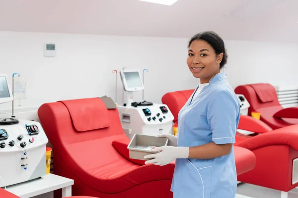 Trabajador sanitario multirracial en uniforme azul y guantes de látex sosteniendo bandeja médica cerca de máquinas de transfusión y cómodas sillas médicas en el centro de donación de sangre - foto de stock
