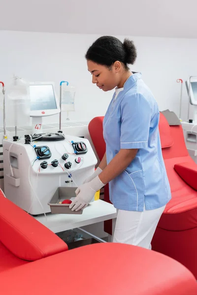 Side view of multiracial nurse in uniform and latex gloves holding medical tray with rubber ball near automated transfusion machine and comfortable medical chairs in blood donation center — Stock Photo