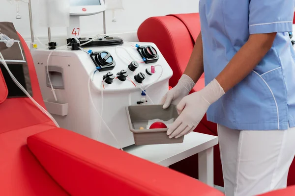Cropped view of multiracial nurse in blue uniform and latex gloves holding medical tray with rubber ball near transfusion machine in blood donation center — Stock Photo