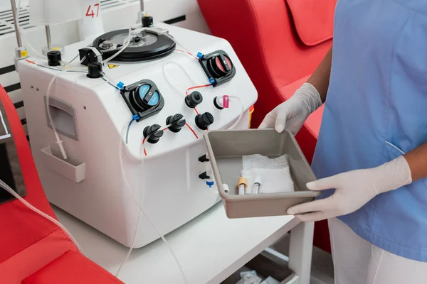 Visão parcial do trabalhador de saúde multirracial em luvas uniformes e látex segurando bandeja médica com tubos de ensaio perto da máquina de transfusão no centro de doação de sangue — Fotografia de Stock