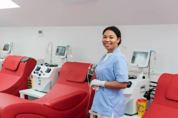 Alegre enfermeira multirracial em uniforme azul e luvas de látex olhando para a câmera perto de cadeiras médicas e máquinas de transfusão com monitores no centro de doação de sangue — Fotografia de Stock