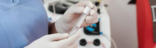 Vista parcial del trabajador sanitario multirracial en uniforme azul y guantes de látex que sostienen el tubo de ensayo estéril cerca de la máquina de transfusión de sangre sobre fondo borroso, pancarta - foto de stock