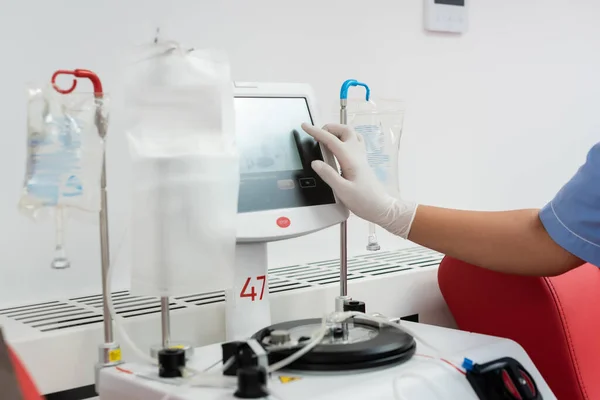 Vista recortada de enfermera multirracial en guante de látex operando máquina de transfusión con pantalla táctil cerca de soportes de goteo con bolsas de infusión en el centro de donación de sangre - foto de stock