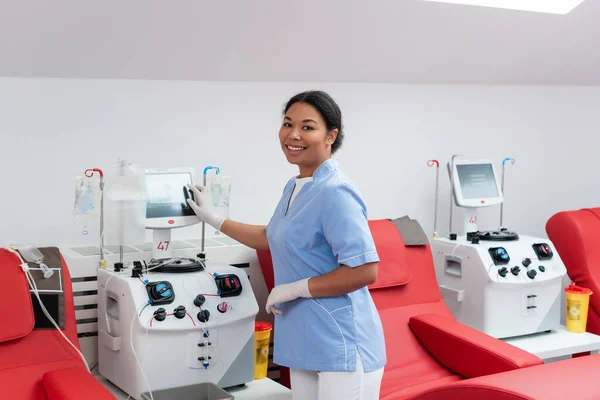 Joven enfermera multirracial en uniforme y guantes de látex operando máquina de transfusión y sonriendo a la cámara cerca de sillas médicas en el centro de donación de sangre - foto de stock