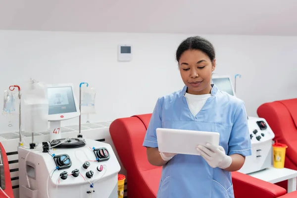 Trabajador sanitario multirracial en uniforme azul y guantes de látex usando tableta digital cerca de sillas médicas y máquinas de transfusión en el centro de donación de sangre - foto de stock
