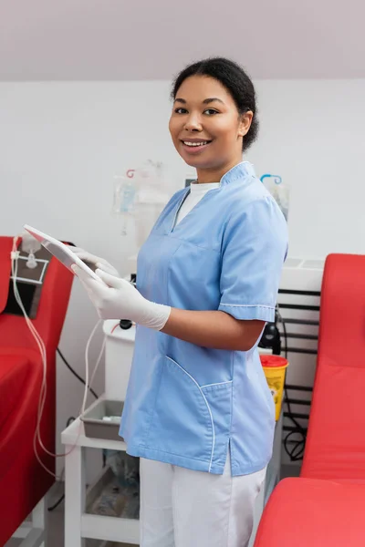 Радостный мультирасовый медицинский работник в форме и латексных перчатках, держащий цифровой планшет и смотрящий на камеру возле медицинских стульев и автоматизированный аппарат для переливания крови в центре донорства — стоковое фото