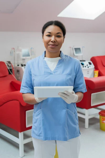 Zufriedene multiethnische Krankenschwester in blauer Uniform und Latexhandschuhen mit digitalem Tablet und Blick in die Kamera in der Nähe von Medizinstühlen und Transfusionsgeräten auf verschwommenem Hintergrund im Blutspendezentrum — Stockfoto