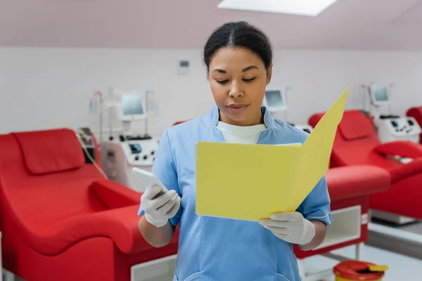 Медицинский работник в синей форме и латексных перчатках, держащий смартфон и читающий медицинские карты возле медицинских стульев и аппаратов для переливания крови в центре донорства — стоковое фото