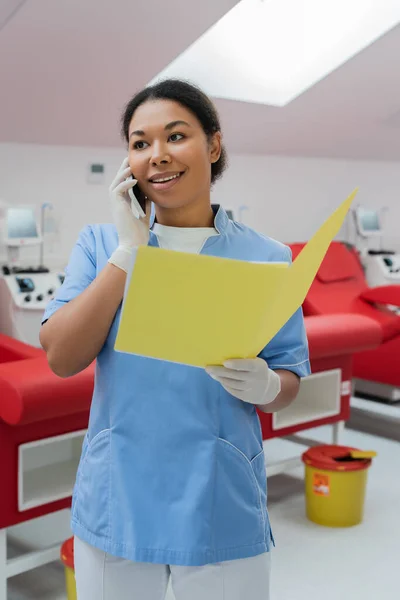 Усміхнена мультирасова медсестра в синій формі і латексні рукавички тримає паперову папку і розмовляє на мобільному телефоні біля розмитих медичних крісел і машин для переливання в центрі донорства крові — стокове фото
