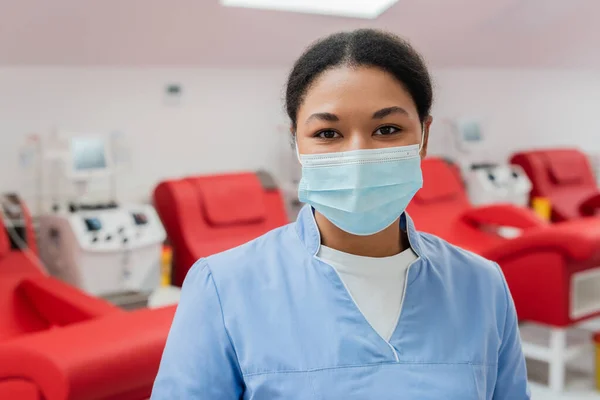 Jeune infirmière multiraciale en uniforme bleu et masque médical regardant la caméra près des chaises médicales et des machines à transfusion sur fond flou à l'hôpital — Photo de stock