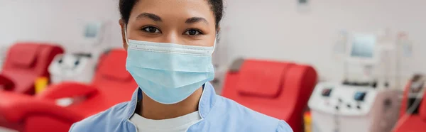 Joven trabajador de la salud multirracial en uniforme azul y máscara médica mirando la cámara cerca de sillas médicas borrosas y máquinas de transfusión en el centro de donación de sangre, pancarta - foto de stock