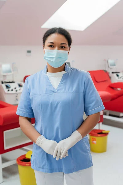 Krankenschwester in blauer Uniform, medizinischer Maske und Latexhandschuhen blickt in die Kamera in der Nähe von Transfusionsmaschinen, medizinischen Stühlen und Mülleimern auf verschwommenem Hintergrund im Blutspendezentrum — Stockfoto