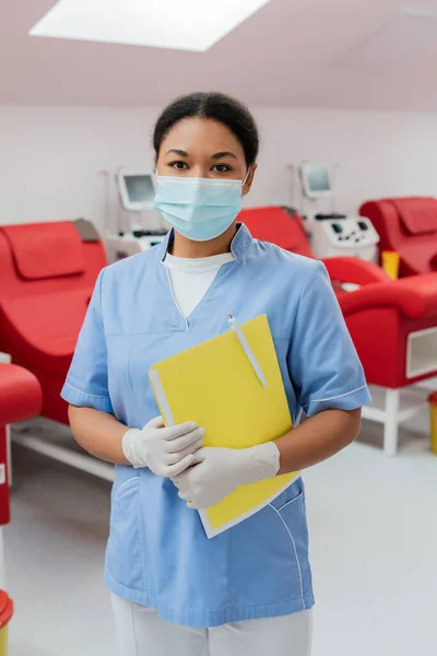 Многорасовый врач в синей униформе, медицинской маске и латексных перчатках, держащий бумажную папку и ручку, глядя на камеру рядом с размытыми машинами для переливания крови и медицинскими стульями в больнице — стоковое фото