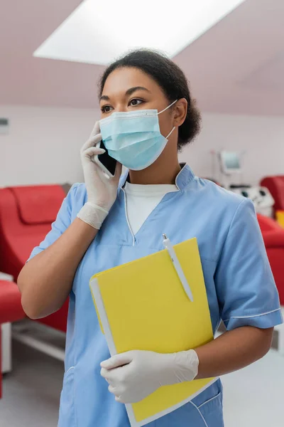 Multirassische Krankenschwester in blauer Uniform, medizinischer Maske und Latex-Handschuhen, die Papiermappe und Stift in der Hand hält, während sie auf dem Smartphone in der Nähe verschwommener medizinischer Stühle in der Bluttransfusionsstation spricht — Stockfoto