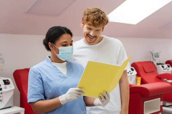 Multiracial nurse in blue uniform, medical mask and latex gloves showing paper folder to smiling redhead man near transfusion machines and blurred medical chairs in blood donation center — Stock Photo