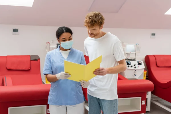 Redhead volunteer and multiracial nurse in uniform, medical mask and latex gloves looking at paper folder near medical chairs and transfusion machines in blood donation center — Stock Photo