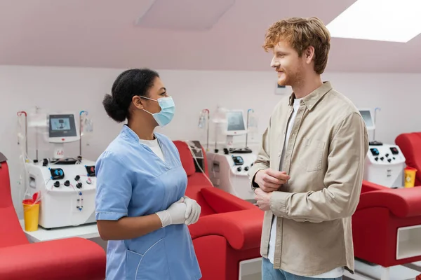 Multiracial nurse in blue uniform, medical mask and latex gloves talking to redhead volunteer near transfusion machines and medical chairs in laboratory — Stock Photo