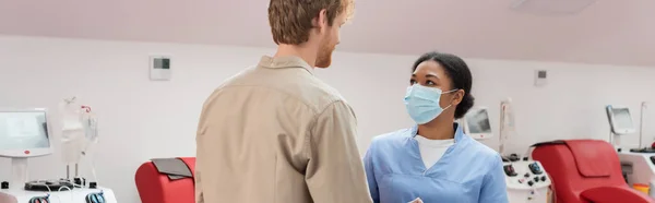 Junge rothaarige Blutspender und multirassische Krankenschwester in blauer Uniform und medizinischer Maske im Gespräch in der Nähe von medizinischen Stühlen und Transfusionsmaschinen im Krankenhaus, Banner — Stockfoto