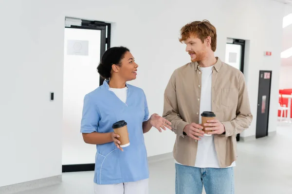Young redhead volunteer and smiling multiracial healthcare worker in uniform holding paper cups while talking in waiting area of blood donation center — Stock Photo