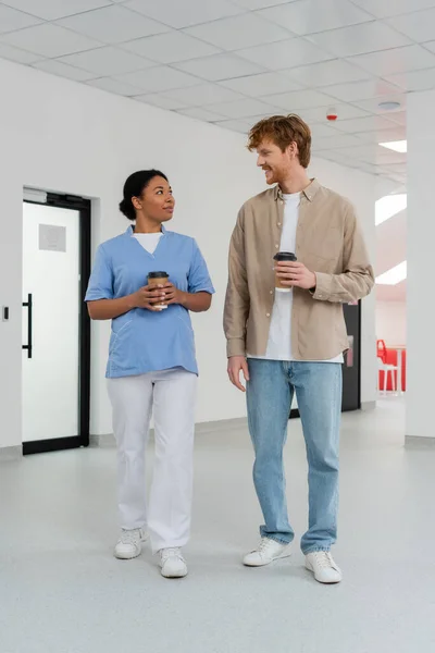 Multiracial nurse in uniform and young, positive redhead volunteer holding paper cups with coffee and talking in waiting area of blood donation center, door on background — Stock Photo