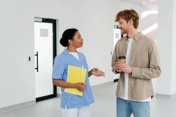 Multiracial nurse in uniform holding paper folder and pen while talking to young redhead volunteer with coffee to go in waiting area of blood transfusion station, door on background — Stock Photo